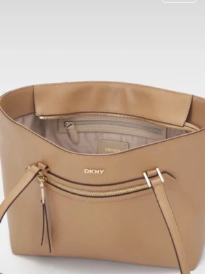 Beige handväska från DKNY - Snygg beige handväska från DKNY i modellen Bryant Ave Medium Tote. Väskan är tillverkad i slätt läder och har guldfärgade detaljer, dragkedja framtill och två axelremmar. Perfekt för dig som vill ha en stilren och rymlig väska med exklusiv känsla.