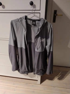 Grå windbreaker från Under Armour - Säljer en snygg tvåtonad grå windbreaker från Under Armour med dragkedja och huva. Windbreaker har en diskret logga på bröstet och är tillverkad i ett mjukt, sportigt material som passar perfekt till chill eller träning.