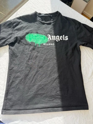 Svart Palm Angels t-shirt Milano - Säljer en svart t-shirt från Palm Angels med Milano-tryck framtill. T-shirten har ett grönt spray paint-inspirerat tryck över loggan och klassisk rund hals. Perfekt för dig som gillar streetwear och vill sticka ut.
