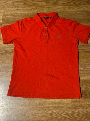 Röd pikétröja från GANT - Klassisk röd pikétröja från GANT med korta ärmar och krage. Tröjan har knappar vid halsen och den ikoniska GANT-loggan broderad på bröstet. Tillverkad i bomull för en skön och luftig känsla.