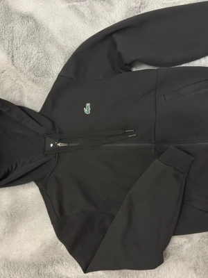 Lacoste kofta - Svart hoodie från Lacoste med dragkedja framtill och klassisk krokodillogga på bröstet. 