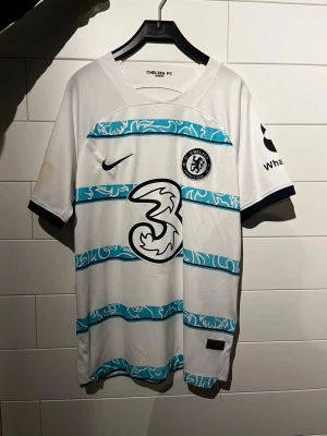 Chelsea FC vit matchtröja Nike - Chelsea FC:s officiella matchtröja i vitt med turkosa ränder och klubbmärke på bröstet. Tröjan har korta ärmar, rund hals och stora tryck på framsidan. Tillverkad i lätt och ventilerande polyester, perfekt för fotboll eller streetwear. Defekt med gul fläck (går att tvätta bort)