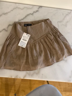 Beige volangkjol från Zara - Supersöt beige kortkjol från Zara med volangdetaljer och luftig känsla. Kjolen har en avslappnad passform och är gjord i ett lätt material som passar perfekt till varmare dagar. Enkel att styla med både sneakers och sandaler. Short under. 