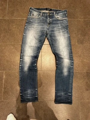 Blå slim fit jeans med slitningar - Säljer ett par blå slim fit jeans med snygga slitningar och tvättade detaljer på framsidan och baksidan. Jeansen har fem fickor, knappgylf och klassisk passform. Perfekta för en avslappnad och trendig stil. Se hål som är i båda fickorna.