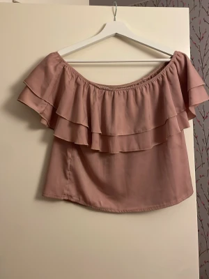 Rosa offshoulder topp med volang - Säljer en ljusrosa offshoulder topp med dubbla volanger. Toppen är ärmlös och har en luftig passform, perfekt för varma dagar. Materialet känns mjukt och lätt, troligen polyester. Snygg att matcha med jeans eller kjol.