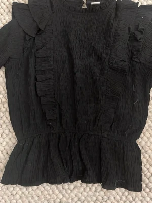 Svart volangblus med struktur - Snygg svart blus med volangdetaljer längs axlar och framtill. Blusen har en krinklad struktur. lätt att både klä upp och ner. Passar perfekt för att skapa ny outfits och passar till mycket. använd bara två eller tre gånger.