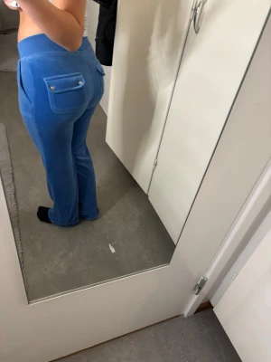Blå juicy mjukisbyxor med fickor - Säljer ett par blå mjukisbyxor i mjuk velour med raka ben och bred resår i midjan. Byxorna har stora fickor fram och bak, bakfickorna har lock och silverfärgade knappar. Perfekta för chill dagar hemma eller när du vill vara extra comfy.