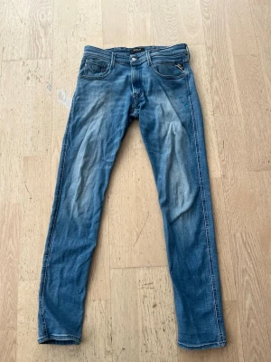 Blå slim fit jeans från Replay - Snygga blå slim fit jeans från Replay med klassisk femficksdesign och ljusa slitningar framtill. Jeansen har en cool tvätt och detaljerade sömmar, samt en liten färgfläck på ena benet. Perfekta för en avslappnad och trendig stil.