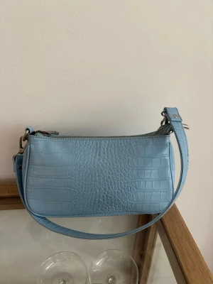 Ljusblå axelväska från Gina Tricot - Snygg ljusblå handväska från Gina Tricot i croco-mönstrat konstläder. Väskan har en avlång form, silverfärgade detaljer och dragkedja 