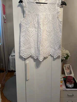 Vit ärmlös broderad blus - Söt vit ärmlös blus med broderade hålmönster och vågig kant. Toppen har rund halsringning och är gjord i ett lätt bomullstyg som passar perfekt till sommaren. Perfekt att styla med jeans eller kjol för en fräsch look.