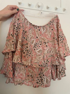 Rosa blommig volangkjol - Supersöt kort kjol i rosa med blommigt mönster och volangdetaljer. Det finns inbyggda shorts i kjolen. Kjolen har en luftig och flowy passform, perfekt för varma dagar. Tillverkad i ett lätt och mjukt material som känns skönt mot huden. 