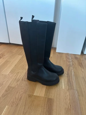 Svarta höga boots i skinnimitation - Säljer ett par svarta höga boots med chunky sula och dragflikar upptill. Stövlarna är i skinnimitation och har en stilren, minimalistisk design som passar till många olika outfits. Grov yttersula för extra grepp och cool look. Storlek 36