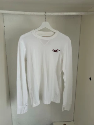 Vit långärmad tröja Hollister - Vit långärmad tröja från Hollister med rund halsringning och det klassiska röda fågel-logotypen broderad på bröstet. Tröjan har ribbade muddar och är tillverkad i ett mjukt, strukturerat bomullsmaterial som känns skönt mot huden.