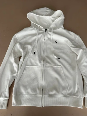 Vit zip hoodie från Polo Ralph Lauren - Klassisk vit hoodie med dragkedja från Polo Ralph Lauren. Tröjan har huva med dragsko, två stora fickor framtill och den ikoniska lilla hästloggan broderad på bröstet. Tillverkad i mjukt bomullsmaterial och har ribbade muddar vid ärmslut och nederkant.