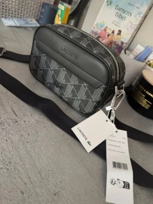 Svart axelväska från Lacoste oanvänd med lappar kvar - Snygg svart axelväska från Lacoste med grafiskt mönster i vitt och silver. Väskan har justerbar axelrem, silverfärgade detaljer och en praktisk ytterficka. Tillverkad i slitstark polyester med dragkedja upptill och diskret Lacoste-logga framtill. Aldrig använd, prislapparna kvar! Alltså inga hål märken osv! Levereras alltid väl inpackat samt snabbt . Hör gärna av dig om du är osäker eller bara har en fråga! Mvh. Sökord: lacoste,väska,snygg,vår,sommar,axelväska,magväska,förvaring. 