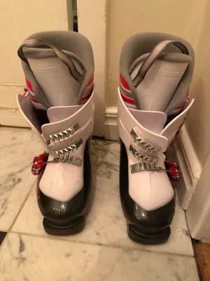 Vita och svarta pjäxor med röda detaljer - Säljer ett par pjäxor i vitt och svart med röda detaljer och metallspännen. Insidan är vadderad i grått tyg för extra komfort. Perfekta för dig som vill ha snygga och funktionella pjäxor till skidbacken. Storlek 36