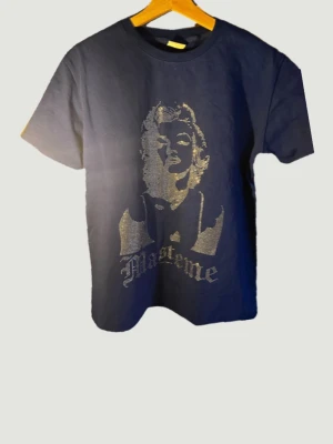 Limiterad T-shirt med glittrigt tryck  - Snygg mörkblå t-shirt med glittrigt tryck av Marilyn Monroe framtill. Klassisk rund halsringning och korta ärmar. Perfekt för dig som vill sticka ut med en cool och trendig look. OBS denna vara går inte längre att köpa och finns endast ett fåtal så passa på medans du kan!