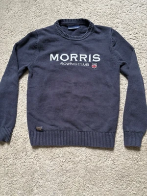 Mörkblå stickad tröja Morris - Mörkblå stickad tröja från Morris med vit text 'MORRIS ROWING CLUB' och en liten broderad detalj på bröstet. Tröjan har rund hals, ribbade muddar och nederkant. Perfekt för dig som gillar klassisk och sportig stil. Storlek S och i bra skick. Tveka inte på att kontakta vid frågor osv.