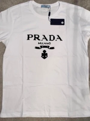 Vit t-shirt från Prada Milano - Vit t-shirt från Prada med svart tryck framtill där det står 'PRADA MILANO' och en ikon under texten. Klassisk rund halsringning och korta ärmar. Perfekt för dig som gillar stilrena och exklusiva plagg.