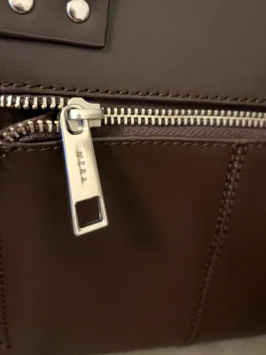 Mörkbrun handväska från Wera - Snygg mörkbrun handväska från Wera i slätt konstläder med silverfärgade metalldetaljer och dragkedja. Väskan har markerade sömmar och en stilren, fyrkantig form. Perfekt för dig som gillar minimalistisk design och vill ha en väska som sticker ut. Nyköpt för 2000kr. Tar endast emot Swish eller kontant vid upphämtning. Skriv i DM vid intresse!💛