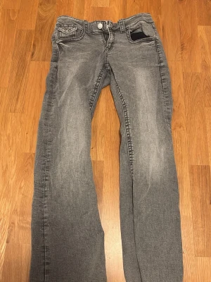 Gråa bootcut jeans med slitningar - Säljer ett par gråa bootcut jeans med snygga slitningar framtill. Jeansen har klassiska fem fickor, knappgylf och detaljerade bakfickor med lock. Perfekta för en avslappnad och trendig look. Materialet är mjukt denim i bomull.