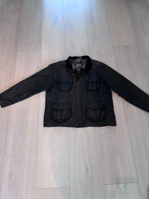 Svart vaxad jacka från Barbour - Klassisk svart vaxad jacka från Barbour med fyra stora fickor framtill och rutigt innerfoder. Jackan har krage i manchester och stängs med både dragkedja och knappar. Perfekt för dig som gillar tidlös brittisk stil. Jackan är ganska sliten och kan behöva vaxas om.