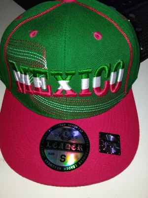 Grön och röd Mexico keps Leader - Snygg snapback-keps i grönt och rött från Leader med stor broderad text 'MEXICO' framtill i vitt, grönt och rött. Platt skärm och detaljerade sömmar. Perfekt för dig som vill sticka ut och visa kärlek till Mexico. Märkets hologramklistermärke kvar på skärmen.
