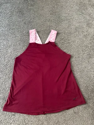 Vinröd linnetopp från Nike Pro - Snygg vinröd linnetopp från Nike Pro med breda rosa axelband där det står 'NIKE PRO' i vitt. Toppen har en lös passform och är perfekt för träning. Materialet känns lätt och andas bra.