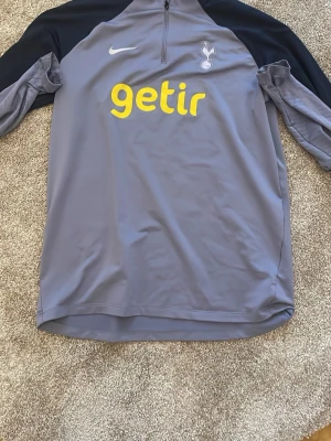 Grå Tottenham träningsjacka Nike - Säljer en grå träningsjacka från Nike med Tottenham Hotspur-logga på bröstet och sponsortryck i gult. Jackan har svarta och grå detaljer på ärmarna samt halv dragkedja framtill. Perfekt för fotbollsträning eller sport.