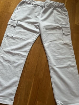 Vita cargobyxor med fickor - Säljer ett par vita cargobyxor med raka ben och stora sidofickor. Byxorna har elastisk midja och är tillverkade i ett mjukt, lätt tyg som känns skönt mot huden. Perfekta för en avslappnad och trendig stil.