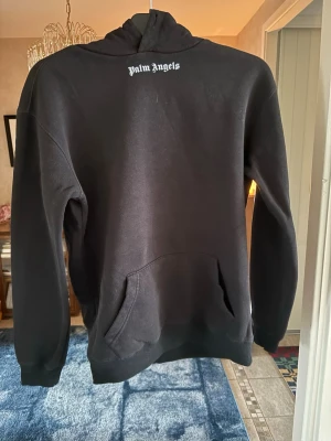 Svart hoodie från Palm Angels - Snygg svart hoodie från Palm Angels med vit logga tryckt på bröstet och stor text på ryggen. Klassisk känguruficka framtill och huva. Perfekt för dig som gillar streetwear och vill ha en stilren men ändå statement look.