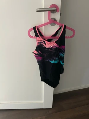 Färgstark baddräkt från SOC - Säljer en snygg baddräkt från SOC med svart bas och färgglada detaljer i rosa, lila och blått. Baddräkten har brottarrygg och ärmlös design, perfekt för simning eller strandhäng. Tillverkad i stretchigt syntetmaterial för bästa komfort.