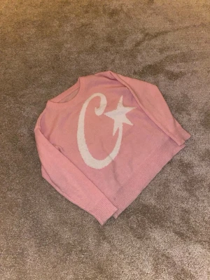 Rosa stickad tröja med stjärnmotiv - Säljer en söt rosa stickad tröja med ett stort vitt motiv av en stjärna och en böjd linje på framsidan. Tröjan har rund halsringning, långa ärmar och ribbade muddar. Perfekt för dig som gillar mysiga och trendiga plagg.