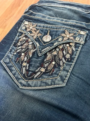 Miss me bootcut jeans - Snygga blå bootcut-jeans från Miss Me med broderade detaljer på bakfickorna. Jeansen har kontrastsömmar och dekorativa nitar, vilket ger en unik och trendig look. Perfekta för dig som vill sticka ut med coola detaljer. Jeansens innerbenslängd är 83cm. Säljer p.g.a dom inte var i min storlek och har därför inga bilder hur dom ser ut på- helt oanvända.