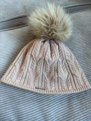 Stickad mössa PaMaMi - Säljer en stickad beige/ljusrosa mössa från PaMaMi med flätmönster och en stor, fluffig pälsboll på toppen. Mössan har en metallogo framtill och är gjord i mjukt material som håller dig varm och snygg under kalla dagar.❄️✨💗💗