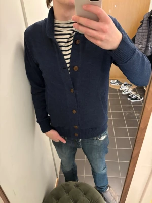 Mörkblå kofta med knappar - Snygg mörkblå kofta med ribbad krage och muddar. Koftan har träknappar framtill och två fickor på sidorna. Perfekt att slänga över en t-shirt för en chill look. Materialet känns mjukt och bekvämt, troligen bomull eller bomullsblandning.