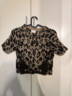 Leopardmönstrad kortärmad kofta\t-shirt - Säljer en kortärmad kofta/t-shirt med leopardmönster i beige och svart. Koftan har rund halsringning, knappar framtill och är i ett mjukt, lurvigt material som känns riktigt cozy. Perfekt för dig som vill sticka ut med ett djurmönster i garderoben.