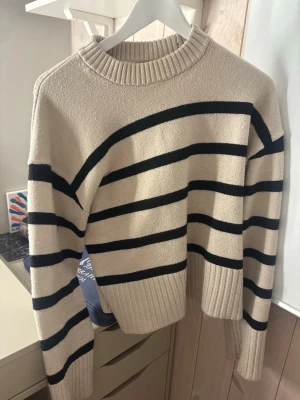 Beige stickad tröja med svarta ränder - Snygg beige stickad tröja med breda svarta horisontella ränder. Tröjan har en ribbad rund halsringning samt ribbade muddar vid ärmslut och nederkant. Perfekt för dig som gillar klassisk och enkel stil.