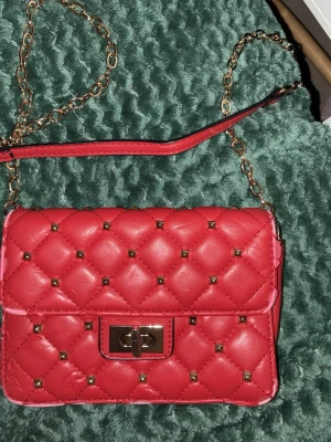 Valentino liknandeRöd quiltad axelväska med nitar - Trendig röd axelväska i quiltat konstläder med små guldfärgade nitar och kedjerem. Väskan har ett snyggt vridlås i guld och en stilren, rektangulär form. Perfekt accessoar för att lyfta din outfit. Från Gina!!