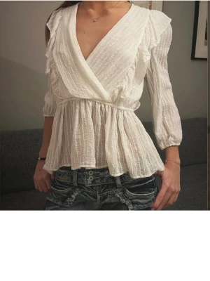 Vit omlottblus med volanger - Söt vit omlottblus med volangdetaljer längs axlarna och v-ringning. Blusen har trekvartsärm och är tillverkad i ett lätt, strukturerat tyg som ger en luftig känsla. Perfekt att styla med jeans eller kjol för en trendig look.