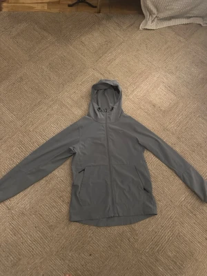Grå vindjacka med huva från Nike - Snygg grå vindjacka från Nike med dragkedja hela vägen, två stora fickor framtill och justerbar huva med snörning. Jackan har en minimalistisk design med diskret Nike-logga på bröstet. Perfekt för blåsiga dagar och enkel att matcha med olika outfits.