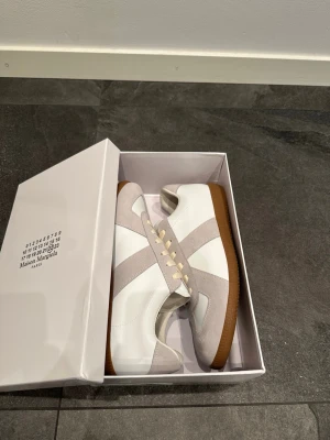 Maison Margiela vita sneakers - Maison Margiela sneakers i vitt läder med ljusgrå mockadetaljer och klassisk brun gummisula. Snygg och stilren design med vita skosnören och diskreta paneler. Perfekt för dig som vill ha en clean look.