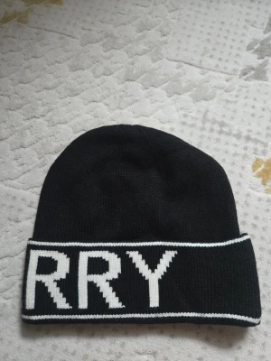 Svart mössa från Burberry - Snygg svart mössa från Burberry med vit logotyptext runt hela uppviket. Tillverkad i mjukt stickat material som håller dig varm och ger en clean streetwear-look. Perfekt accessoar för att lyfta din outfit under kyliga dagar.