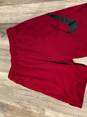 Röda Nike shorts med svart Swoosh - Snygga röda shorts från Nike med stor svart Swoosh-logga på sidan. Shortsen har elastisk midja och sidofickor, perfekta för en sportig look. Tillverkade i ett lätt och bekvämt material som passar bra till träning eller chill.