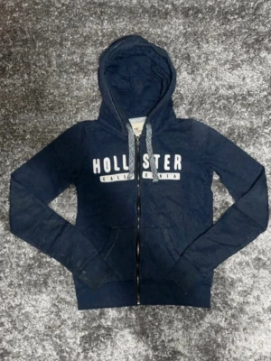 Marinblå hoodie från Hollister - Snygg marinblå hoodie från Hollister med dragkedja och vit logga på bröstet. Hoodien har huva med snören och två fickor framtill. Används inte då den är för liten.