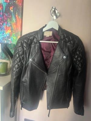 Svart skinnjacka med quiltade axlar - Snygg svart skinnjacka med quiltade detaljer på axlarna och silvriga dragkedjor. Jackan har en cool bikerstil och vinrött innerfoder. Finns inget att anmärka på, mycket fint skick storlek 40 passar en medium