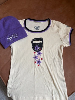 Merch- Vit t-shirt med tryck från Olivia Rodrigo - Cool vit t-shirt från Olivia Rodrigo med svart tryck av en mun och lila stjärnor med texten 'GUTS' samt röda stjärnor. Lila och svart kant vid hals och ärmslut. Perfekt för dig som gillar unik och trendig merch. Den är som en Svmen i tröjan står L