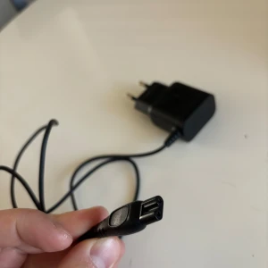 Svart laddare Philips  - En svart laddare med väggadapter. Sladden är tunn och flexibel, och adaptern har två stift för vägguttag. Perfekt för att ladda mindre elektronikprylar. Märket syns inte tydligt på bilderna.