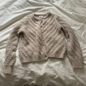 Beige stickad kofta från only - Mysig beige stickad kofta från only med diagonalt mönster och rund halsringning. Koftan har långa ärmar, ribbade muddar och knäppning framtill med små knappar. Perfekt för lager på lager och en avslappnad look.