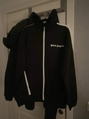 Palm Angels svart ziptröja - Säljer en svart ziptröja från Palm Angels med vita detaljer. Tröjan har vit dragkedja, vita ränder längs ärmarna och logga på bröstet. Hög krage och sportig look, perfekt för dig som gillar streetwear.